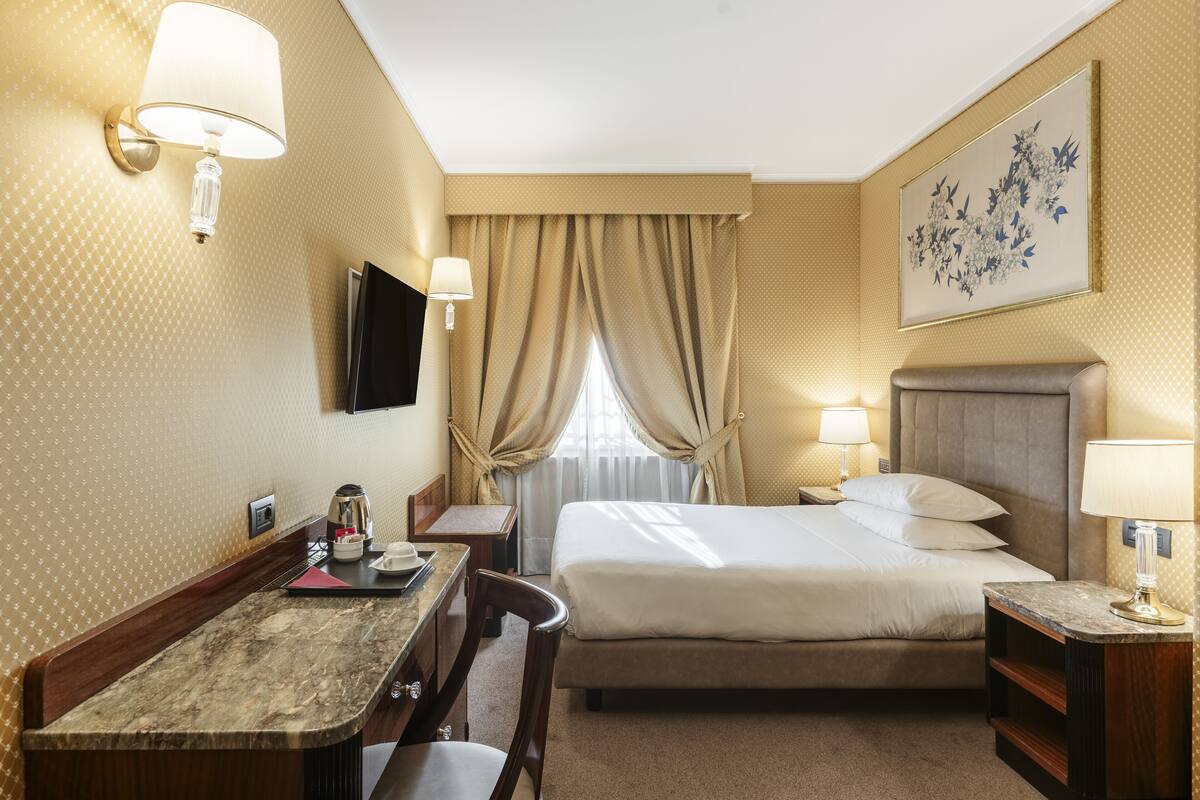 A hotel room with a bed, bedside tables, a desk, a chair, a TV, and a window with curtains.