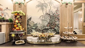A buffet setup with various fruits and desserts, a decorative wall with a tropical mural, and a chandelier. - Doria Grand Hotel (Milan)