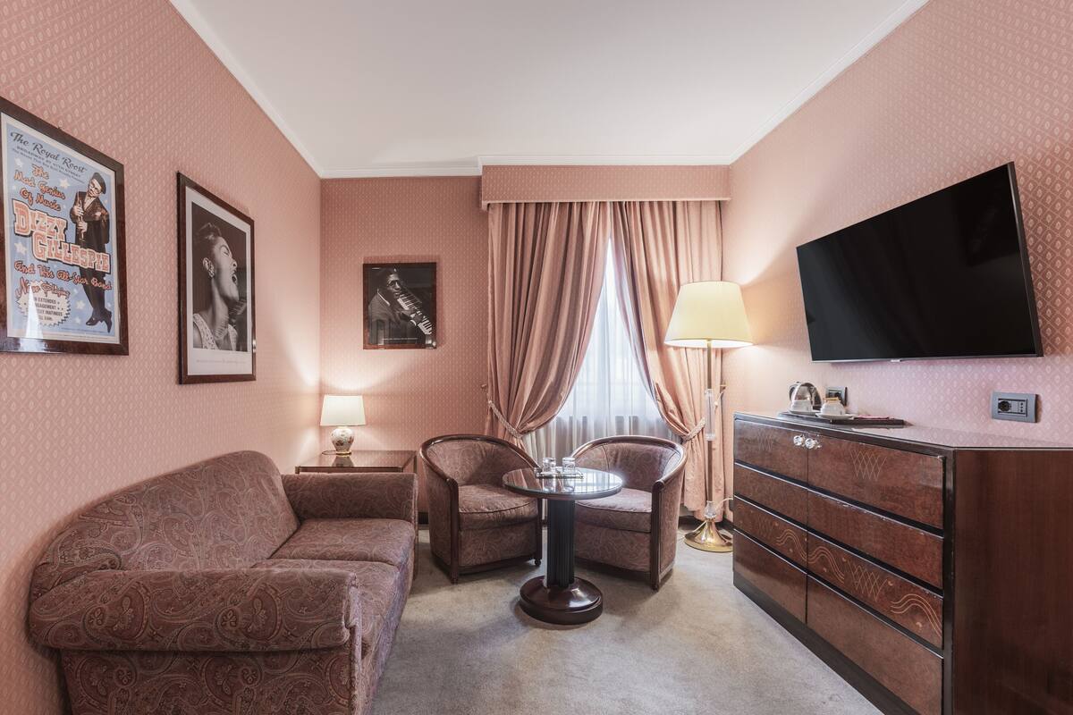 A hotel room with a sofa, two armchairs, a small round table, a TV, a dresser, and curtains.