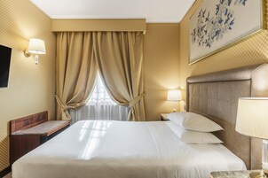 Ένα δωμάτιο ξενοδοχείου με ένα μεγάλο κρεβάτι, ένα ξύλινο παγκάκι, ένα κομοδίνο με ένα φωτιστικό και μια τηλεόραση τοποθετημένη στον τοίχο.