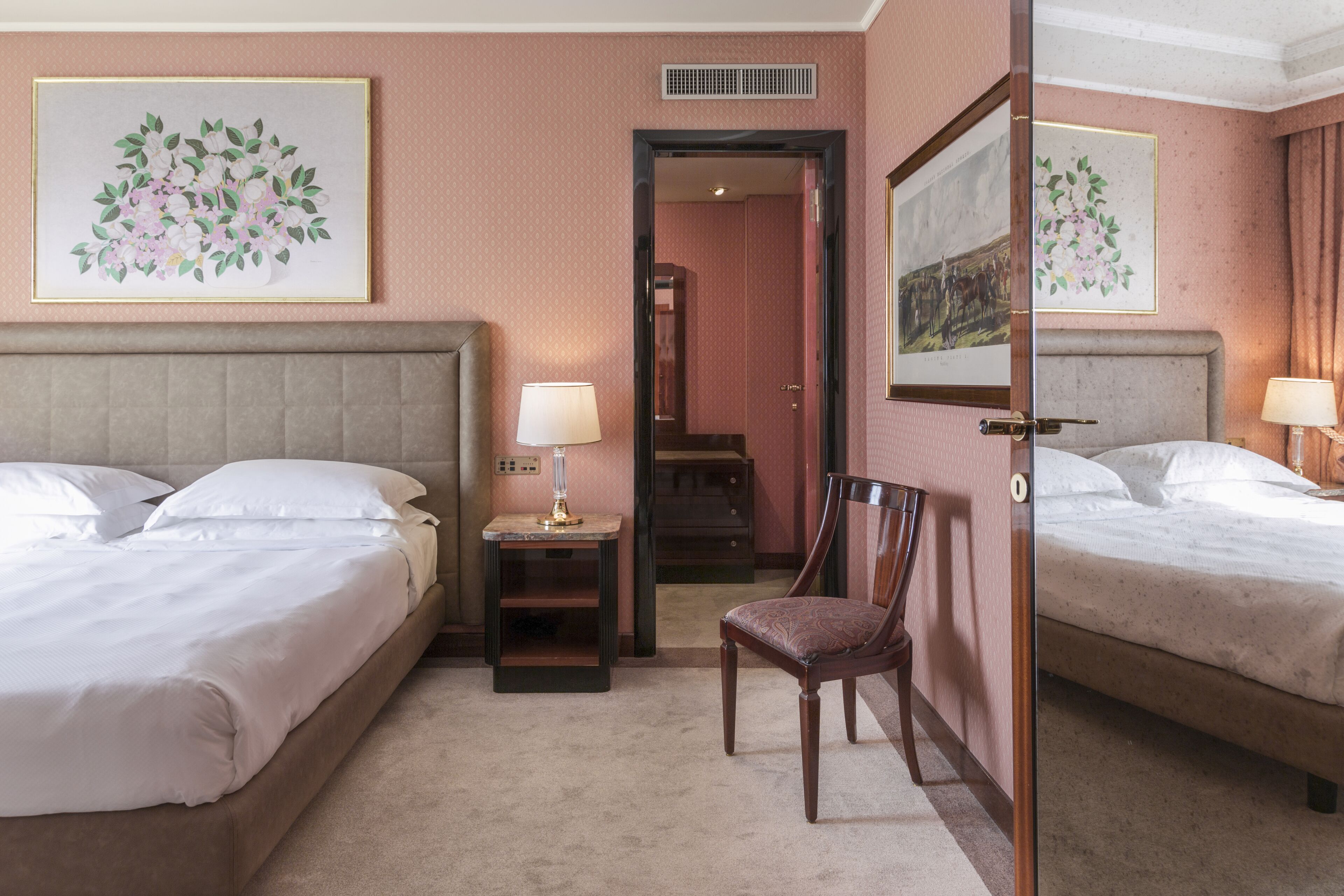 A hotel room with two beds, a chair, a nightstand with a lamp, and framed floral artwork on the walls.
