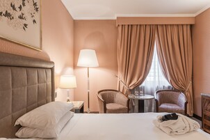 Ένα δωμάτιο ξενοδοχείου με ένα μεγάλο κρεβάτι, δύο καρέκλες, ένα μικρό τραπέζι με ένα φωτιστικό και ένα παράθυρο με κουρτίνες.