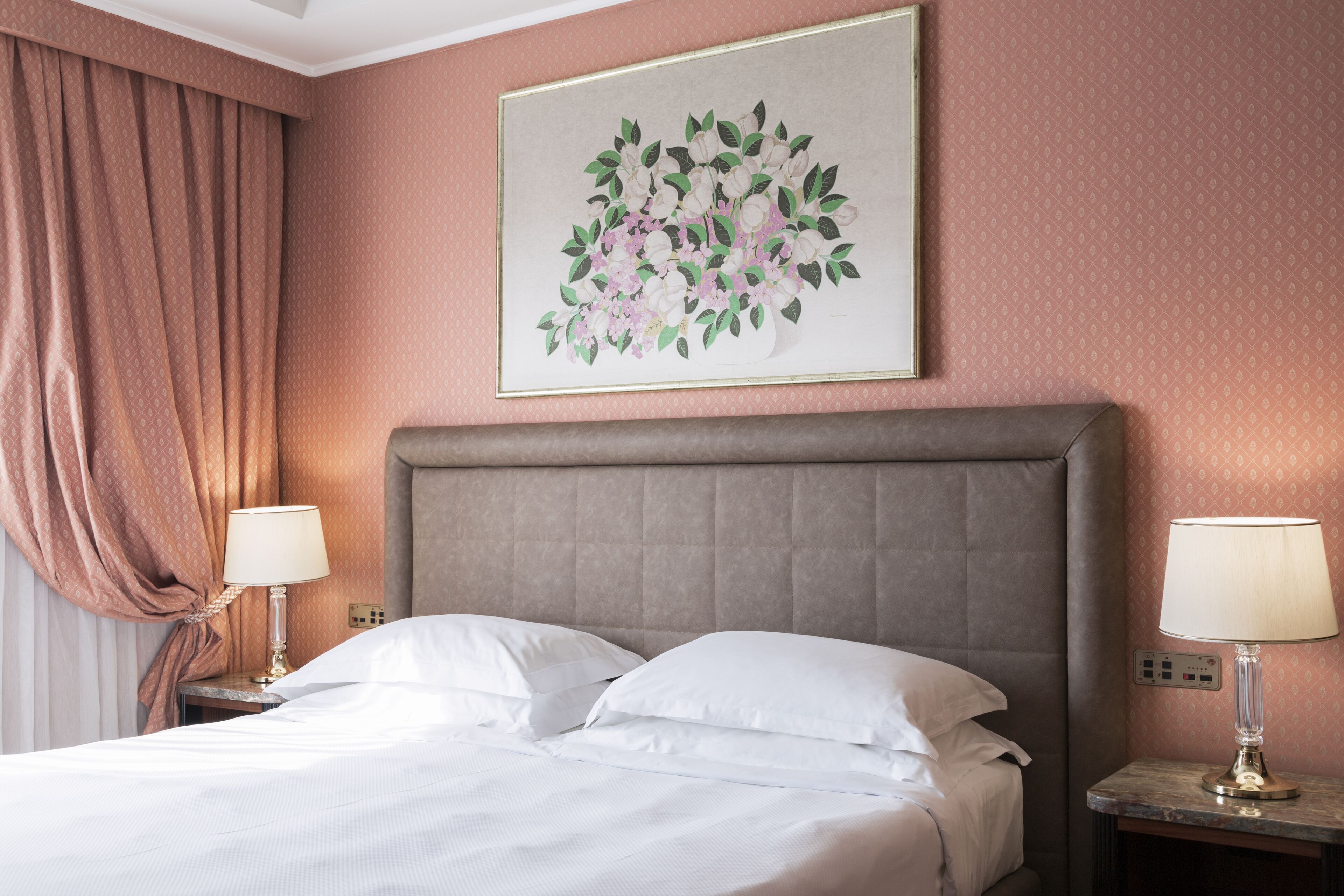 Un letto rifatto con lenzuola bianche, una testiera, due abat-jour e un quadro di fiori appeso alla parete.