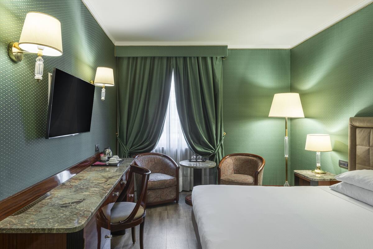 A hotel room with a bed, a desk with a chair, a round table, two armchairs, a TV, and a window with curtains.