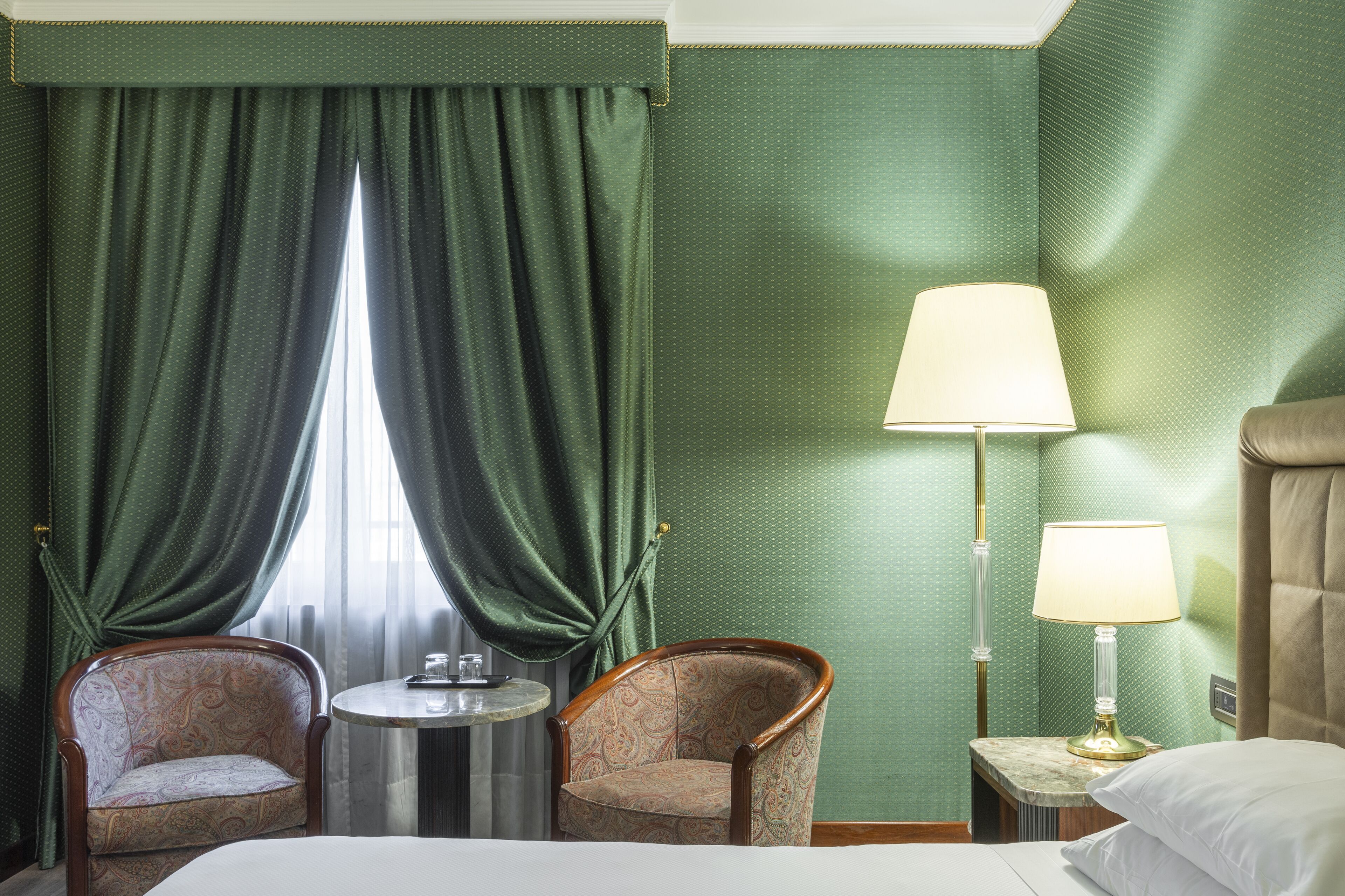 Une chambre d’hôtel avec un lit, deux fauteuils, une petite table ronde, une lampe et une fenêtre avec des rideaux verts.