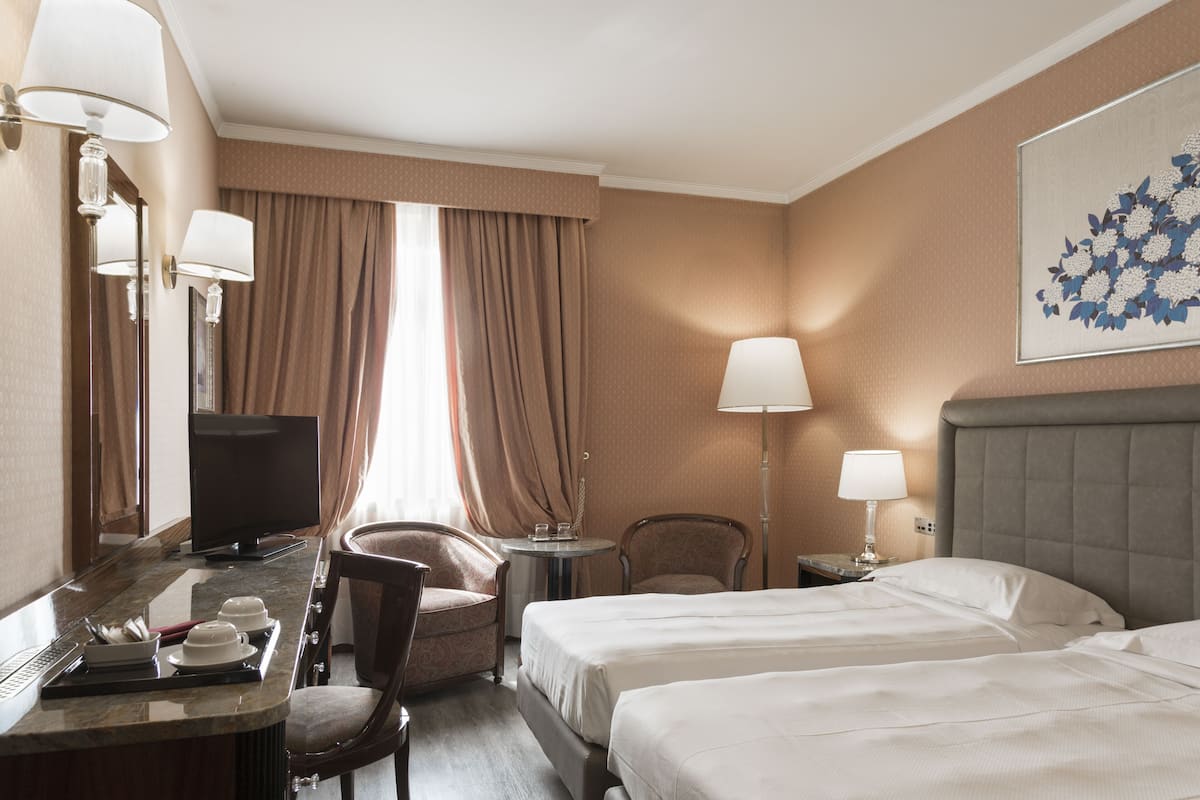 A hotel room with two beds, a desk with a computer, a chair, a small table, and a lamp.