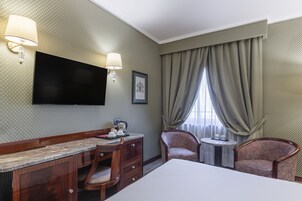 A hotel room with a large bed, a desk with a chair, a small table, a TV mounted on the wall, and a window with curtains.