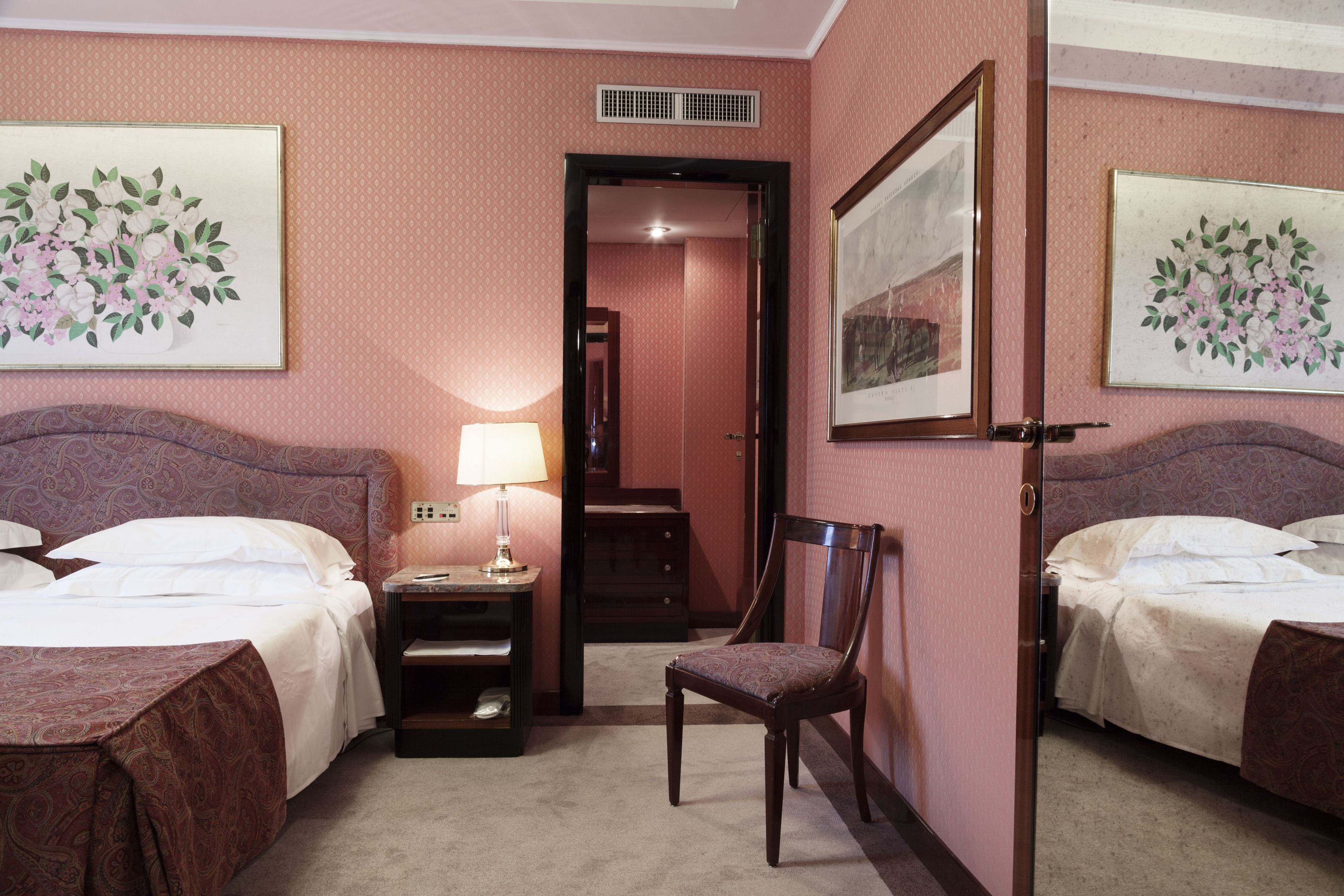 Habitación de hotel con dos camas, una silla, una mesita de noche con lámpara y obras de arte en las paredes.