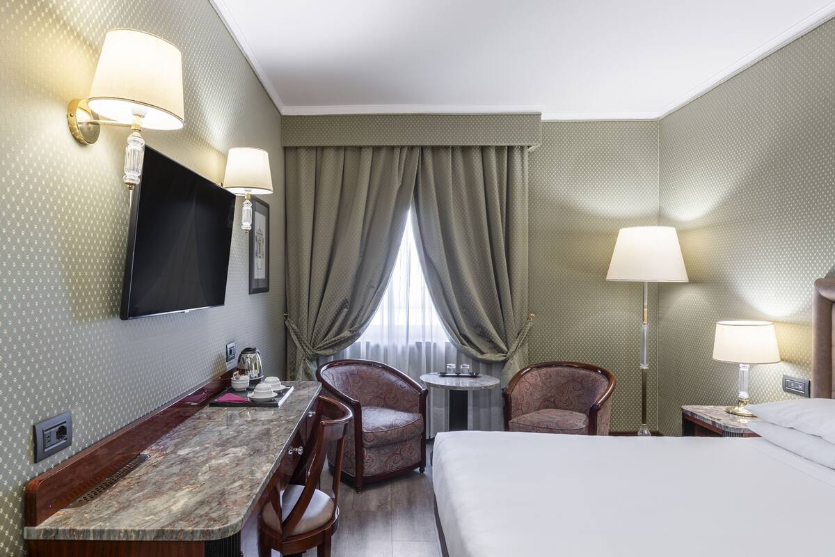 A hotel room with a bed, two armchairs, a desk with a mirror, a television mounted on the wall, and a window with curtains.