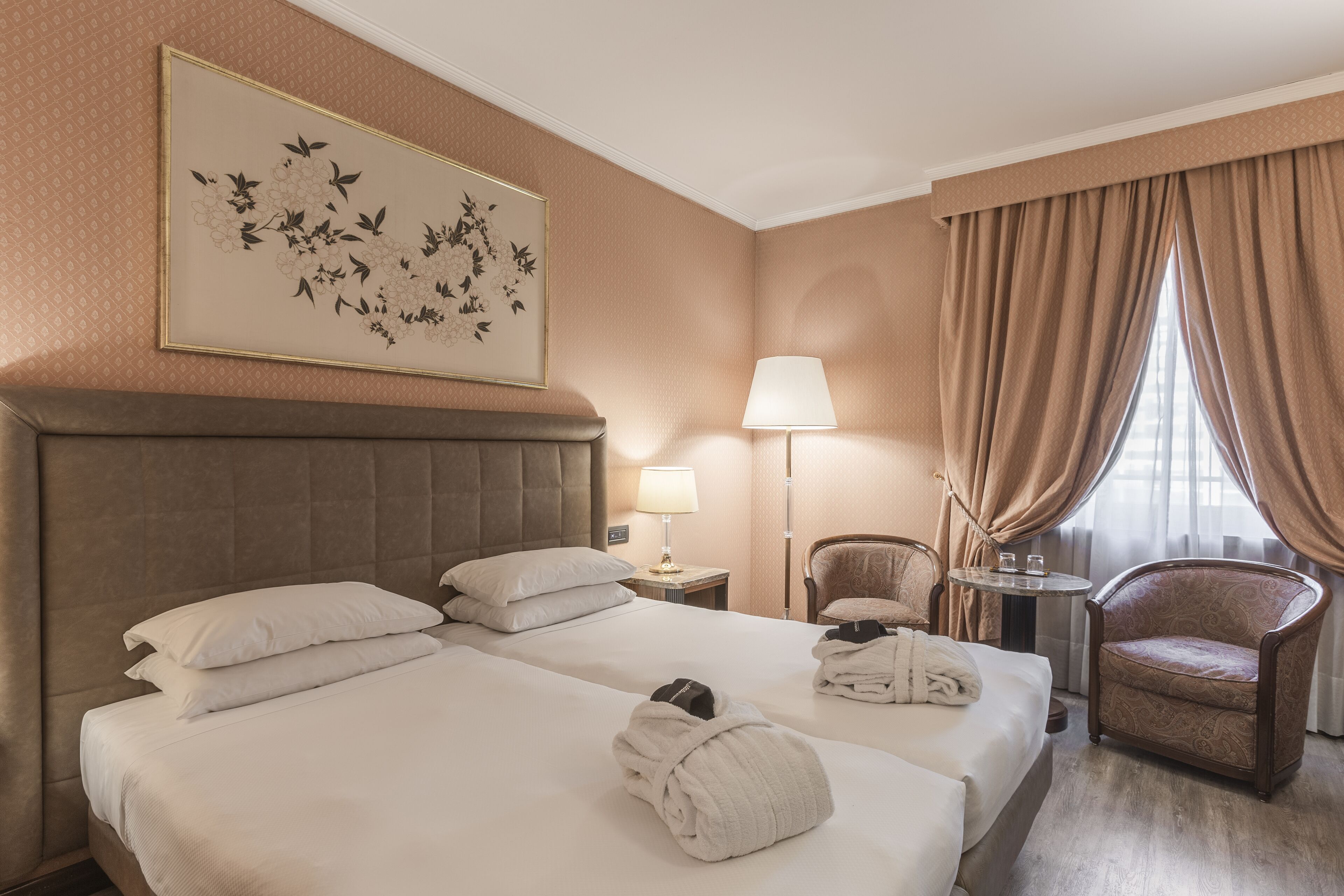 A hotel room with two beds, a headboard, a lamp, a chair, a table, and a window with curtains.