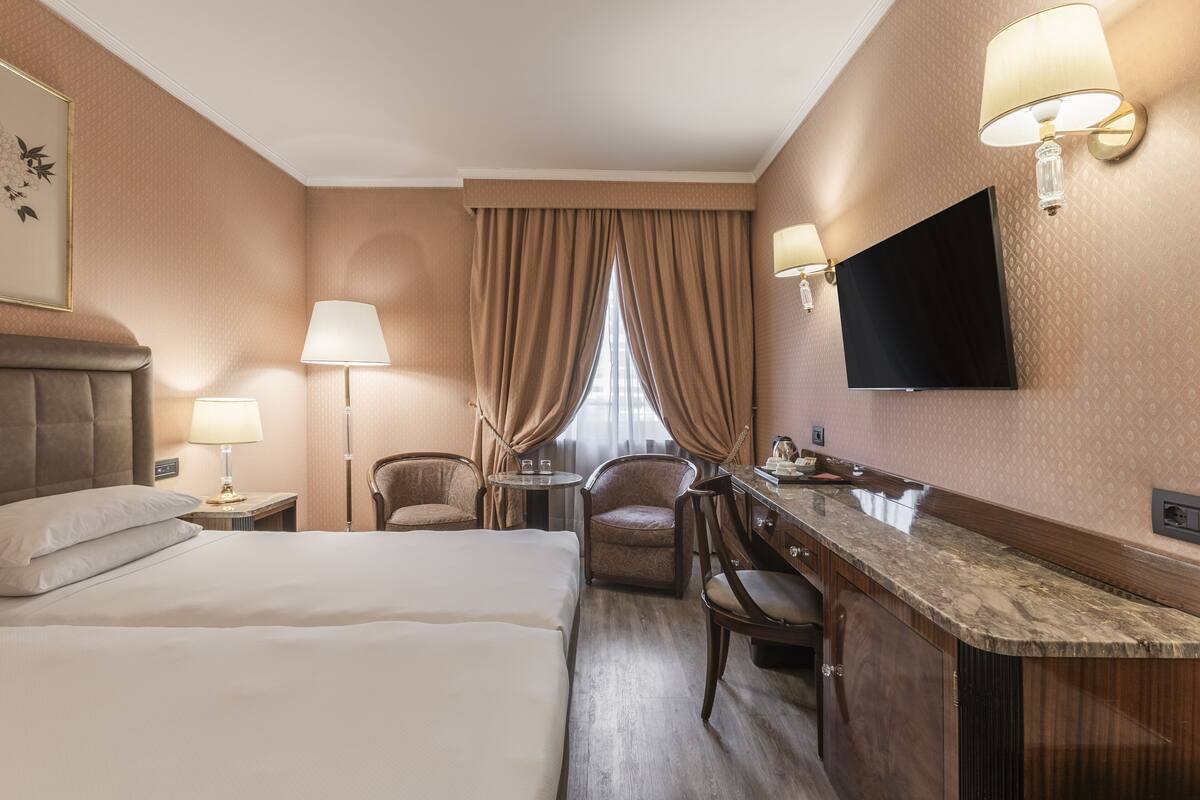 A hotel room with a bed, a desk with a chair, a TV, and a window with curtains.