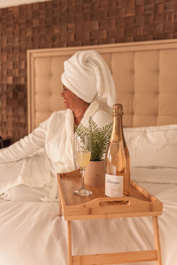 A person in a white robe sitting on a bed with a wooden tray holding a bottle of champagne and a glass.