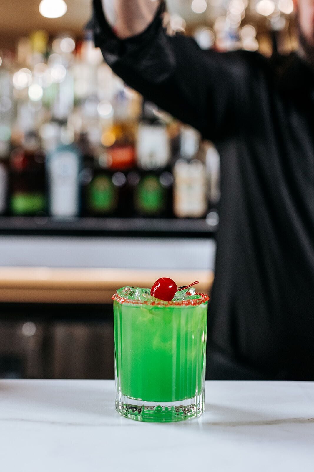 En bardisk med en grön cocktail i ett glas, garnerad med ett körsbär, och en suddig bakgrund med olika flaskor.