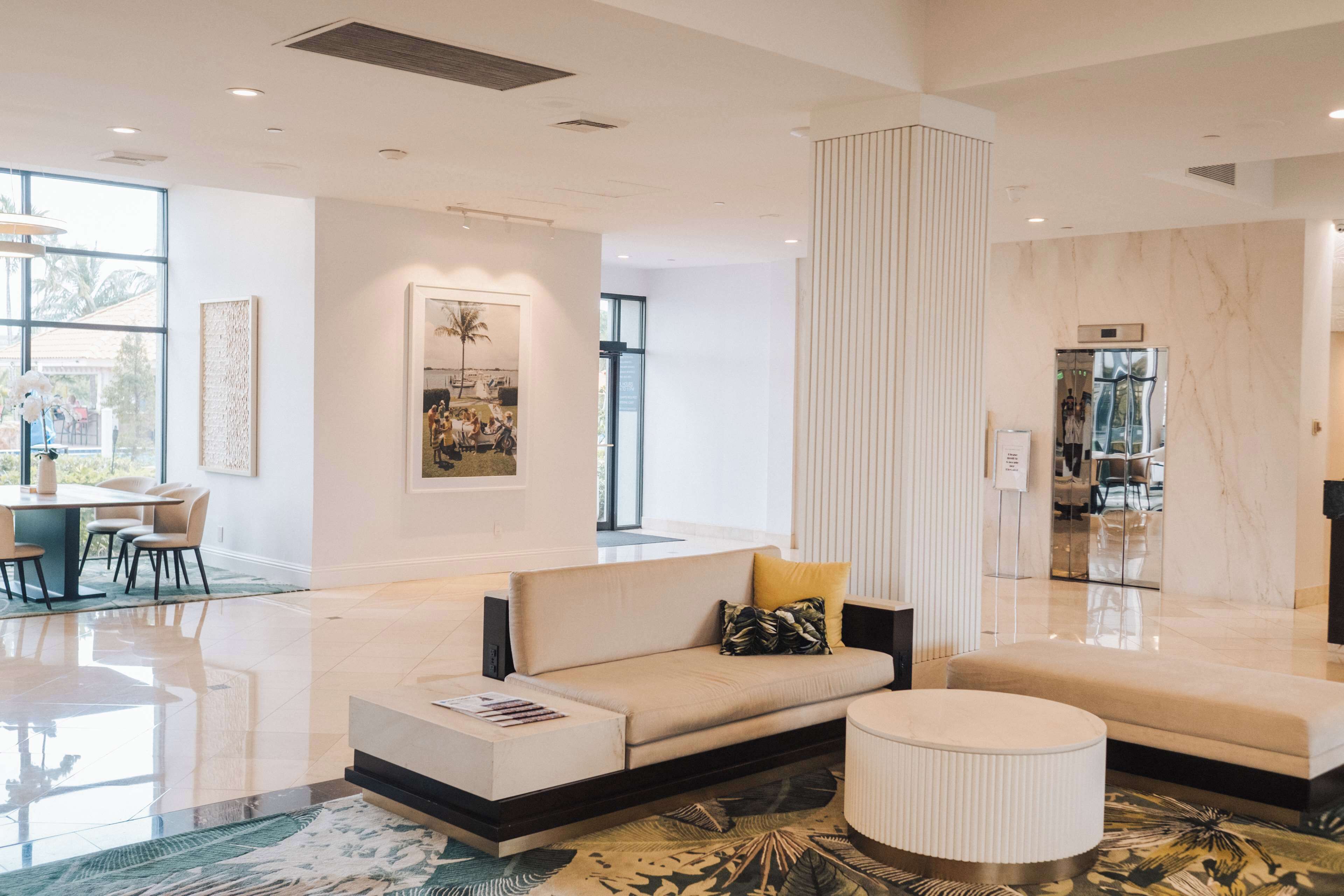 Un moderno vestíbulo de hotel con un sofá, reposapiés y una zona de comedor con sillas y una mesa.