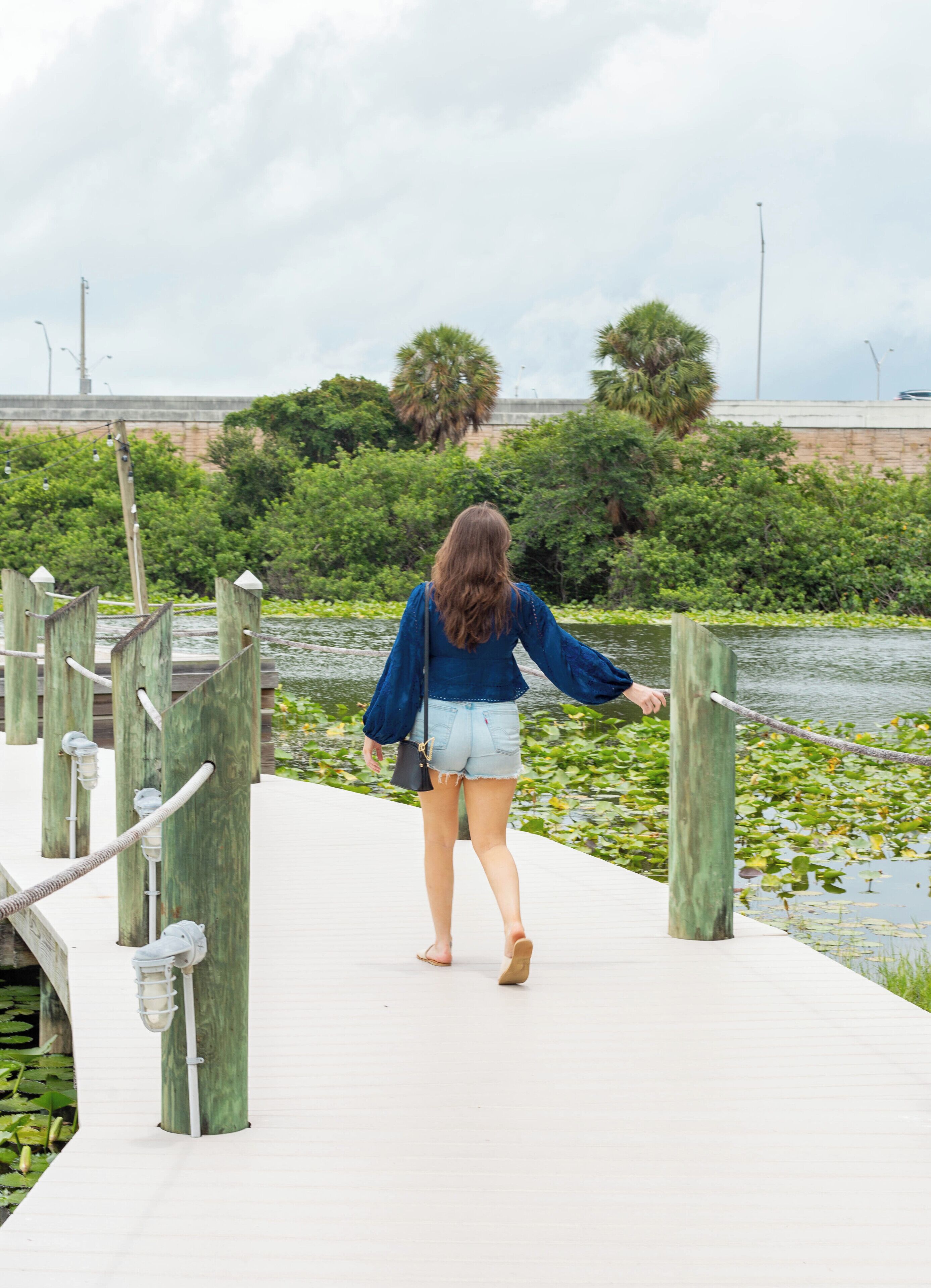 Una mujer caminando sobre un muelle de madera, con un río y follaje al fondo.