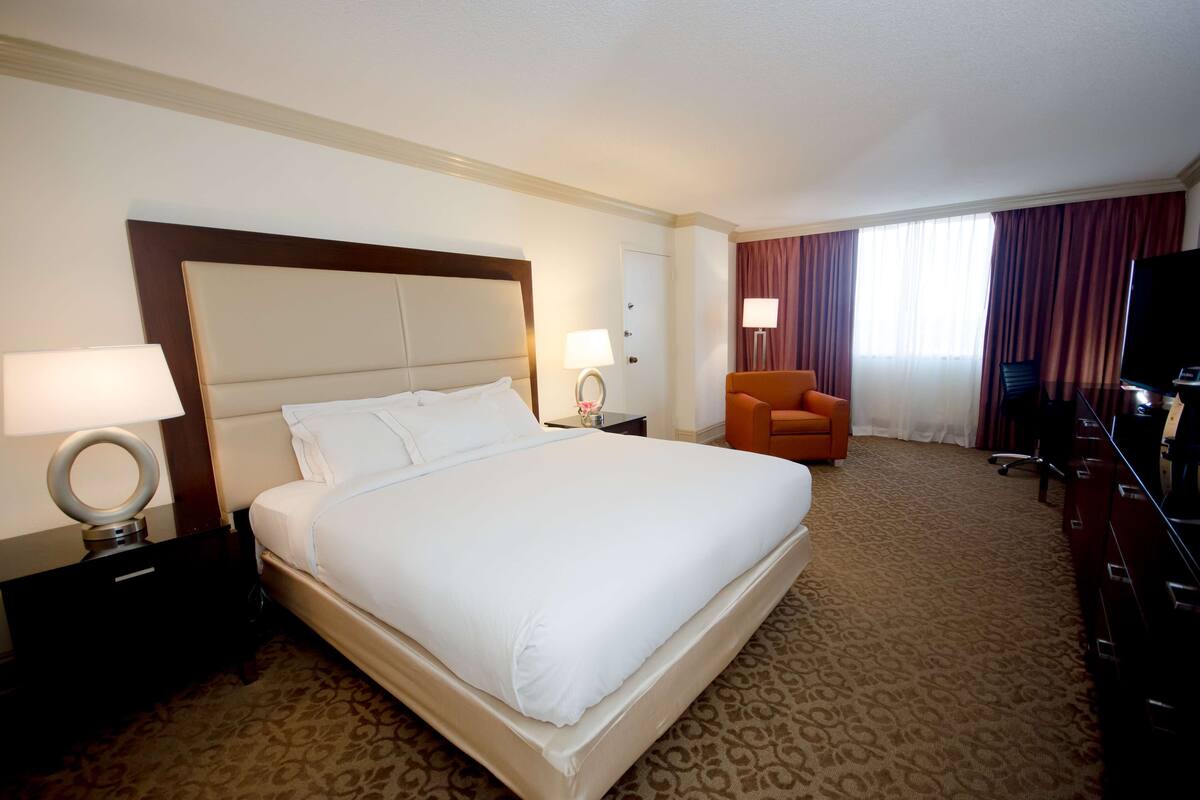 Une chambre d’hôtel avec un grand lit, deux tables de chevet avec des lampes, une chaise, un bureau avec une télévision et une fenêtre avec des rideaux.