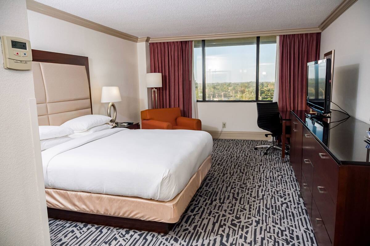 Une chambre d’hôtel avec un grand lit, un bureau avec une télévision, une chaise et une fenêtre avec des rideaux.