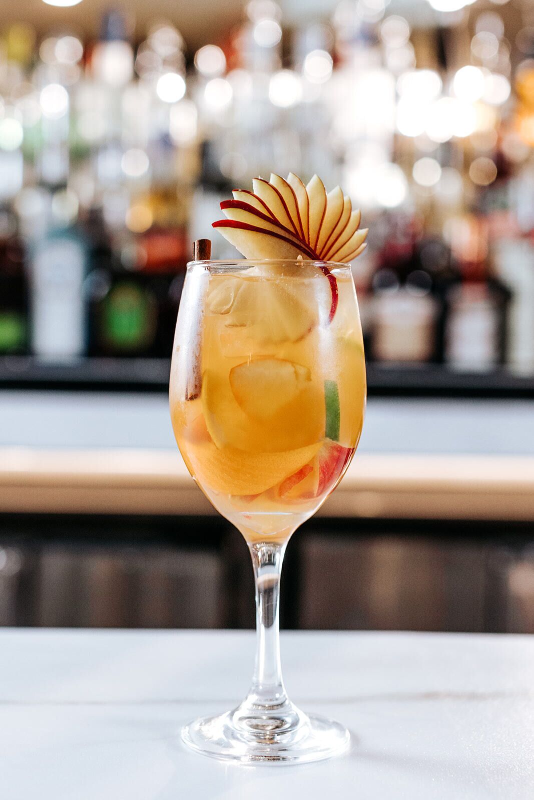A cocktail in a stemmed glass with fruit garnish, ice cubes, and a blurred bar background.