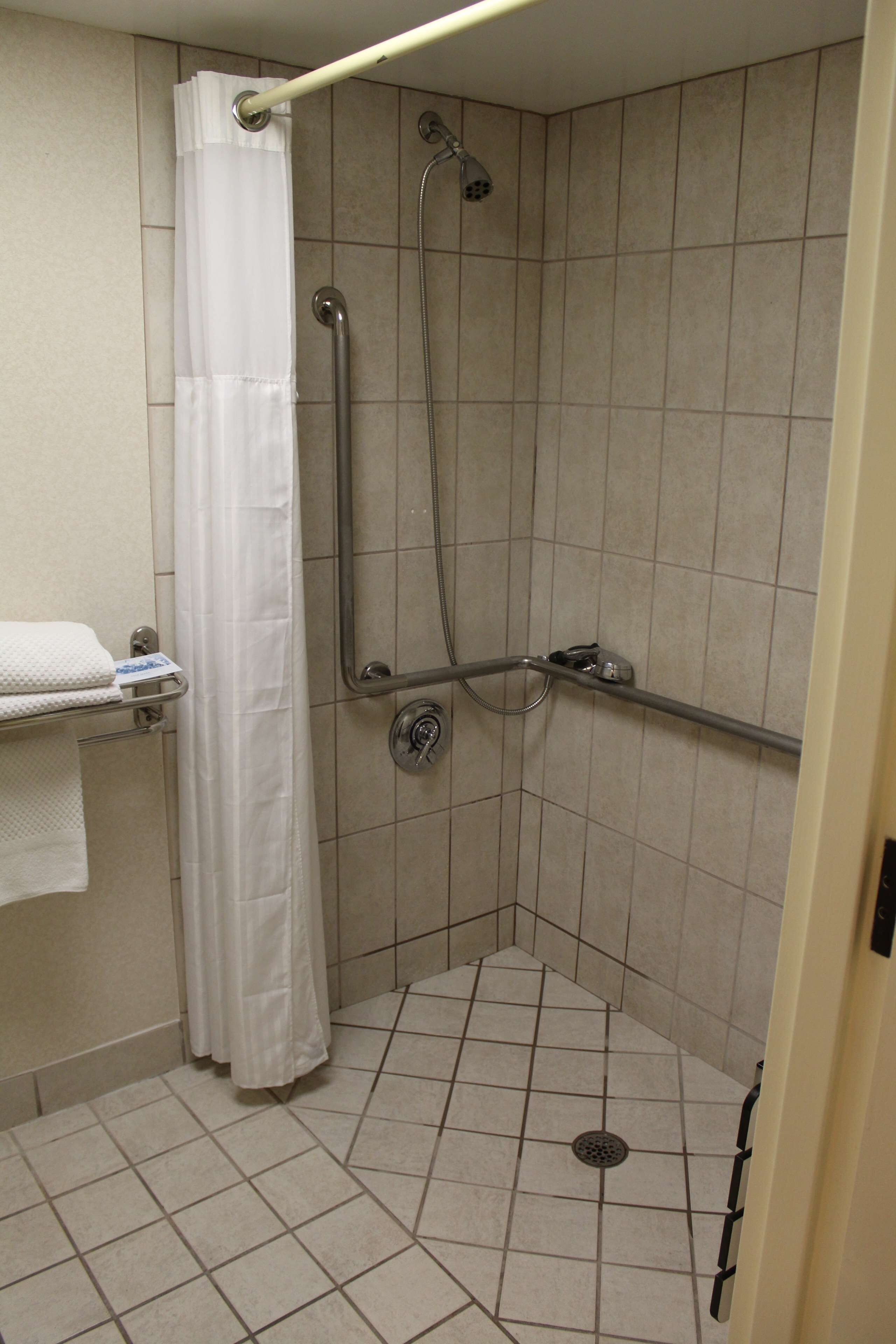Un espacio de ducha pequeño, con azulejos, barra de apoyo, cortina blanca y cabezal de ducha manual.