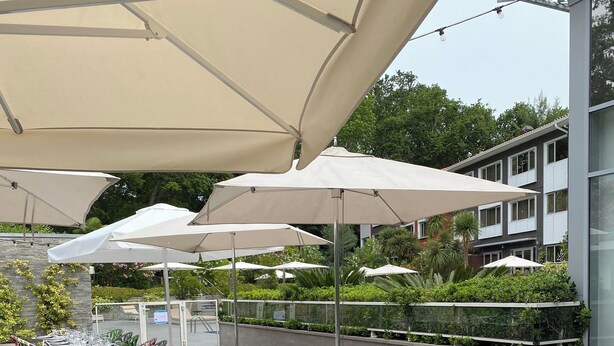 A long outdoor dining area with tables and chairs, large umbrellas, and a view of a building and greenery in the background. - Novotel Resort & Spa Biarritz Anglet (Anglet)