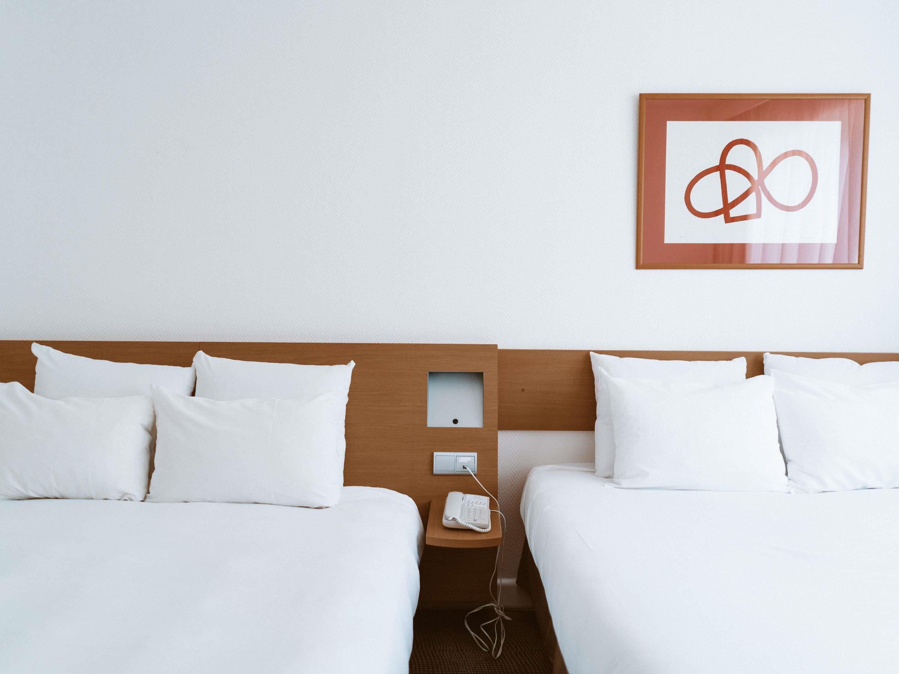 A hotel room with two beds, a wooden headboard, a bedside table with a phone, and a framed abstract artwork on the wall.