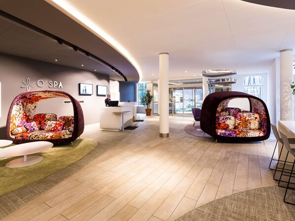 A modern spa reception area with a curved ceiling, a reception desk, and two floral-patterned seating areas. - Novotel Resort & Spa Biarritz Anglet (Anglet)