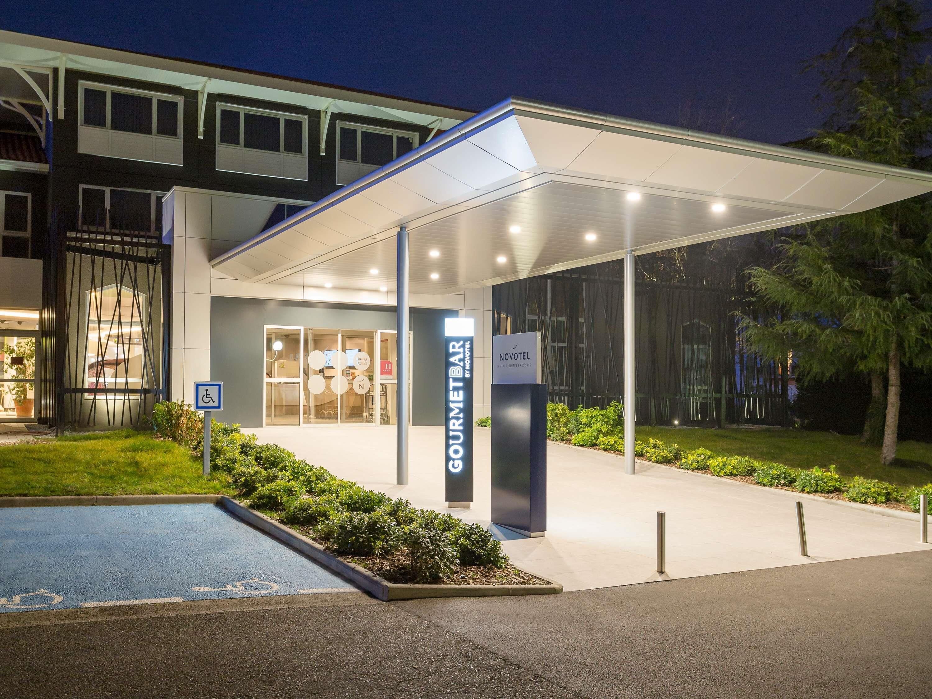 Entrada de un hotel con un corredor cubierto, un cartel con la leyenda 'NOVOTEL' y un espacio de estacionamiento para discapacitados.