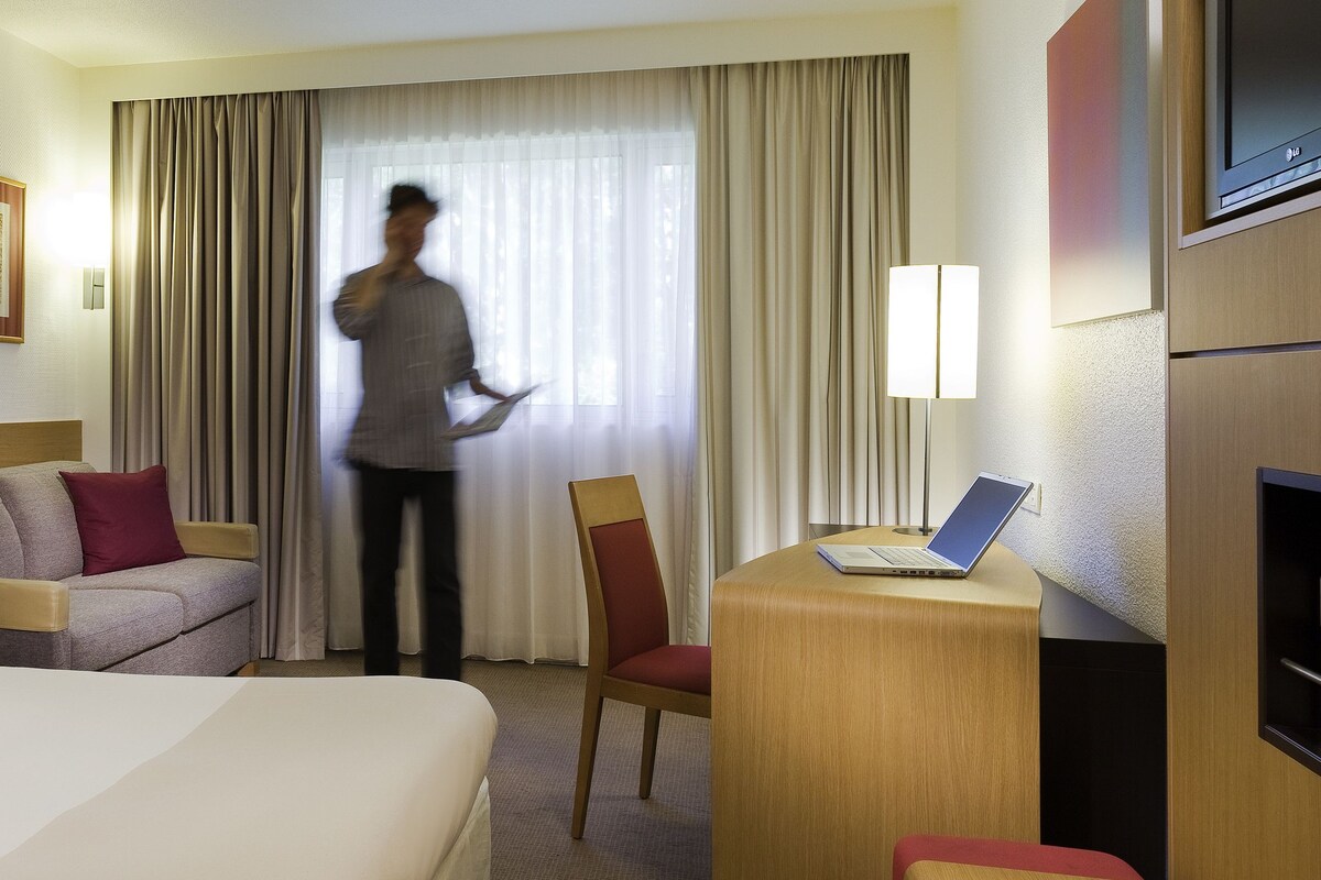 A hotel room with a bed, a desk with a laptop, a chair, a sofa, and a window with curtains.
