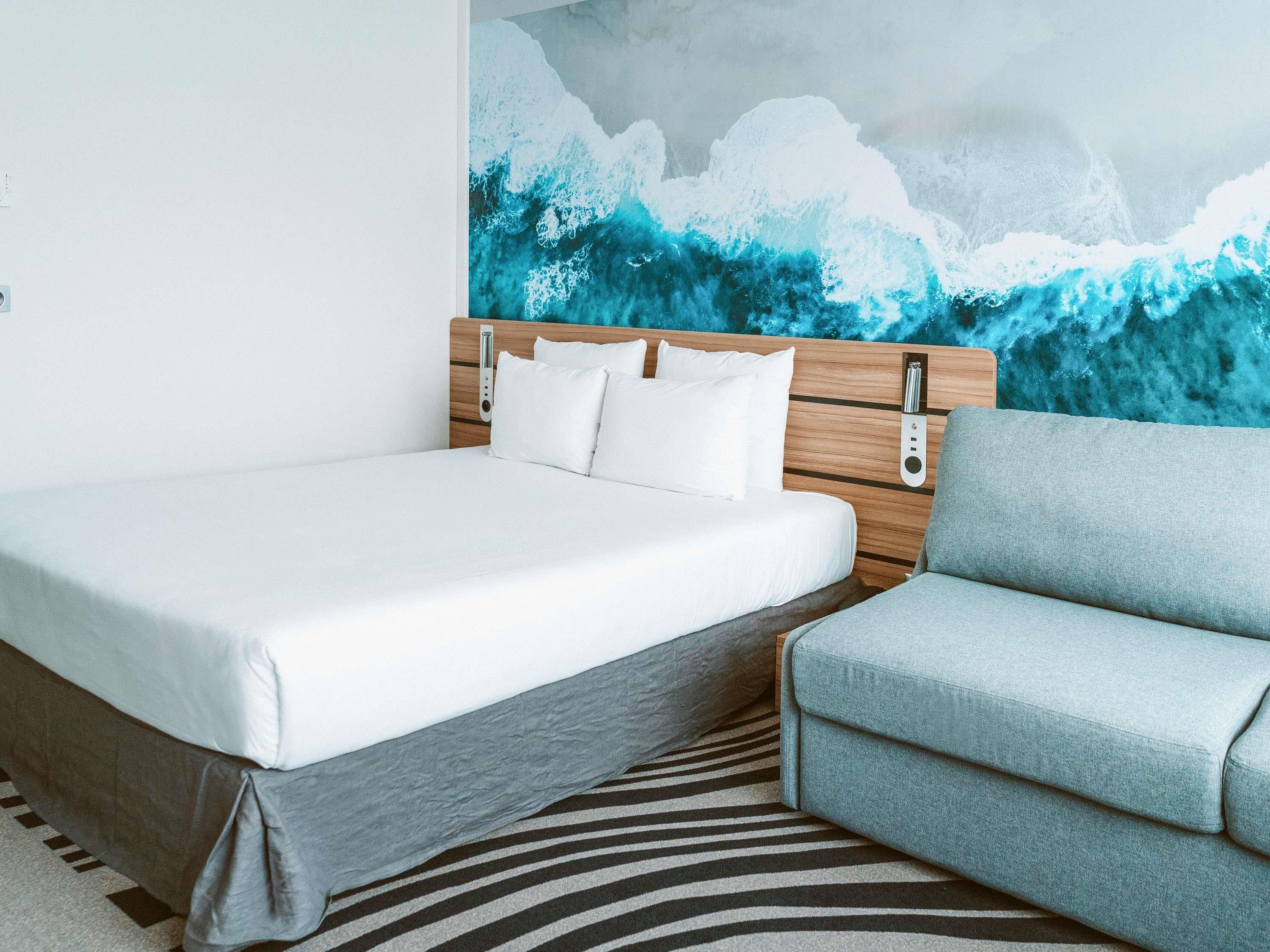 A hotel room with a bed, a grey sofa, a zebra-striped rug, and a wall mural of a wave.