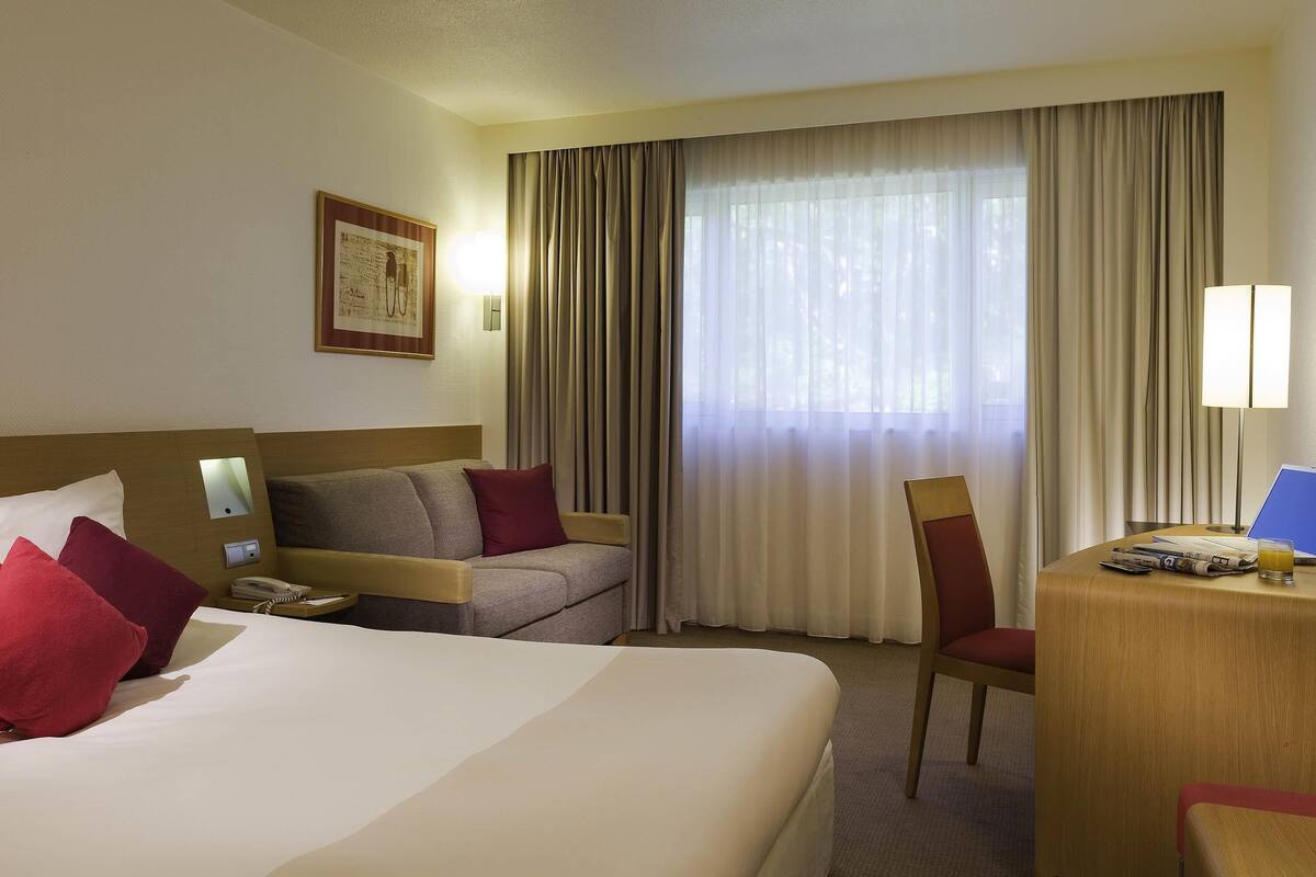 Habitación de hotel con una cama grande, un sofá, un escritorio con lámpara y una ventana con cortinas.