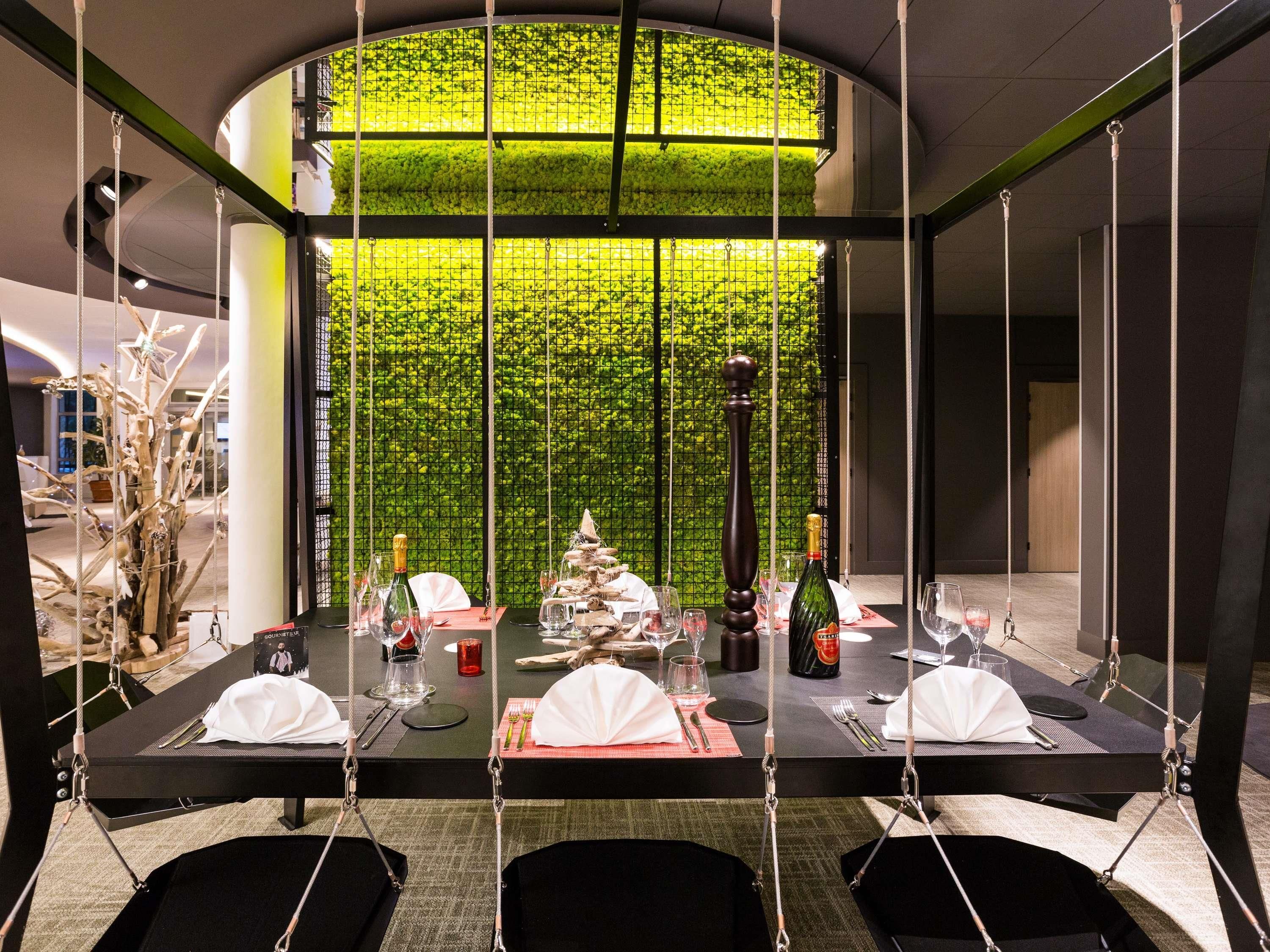 Un comedor con techo alto y pared verde. Las mesas están dispuestas con manteles blancos, copas de vino y botellas.
