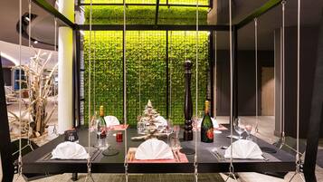Un comedor con techo alto y pared verde. Las mesas están dispuestas con manteles blancos, copas de vino y botellas.