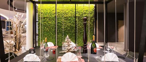 A dining area with a high ceiling and a green wall. Tables are set with white tablecloths, wine glasses, and bottles.