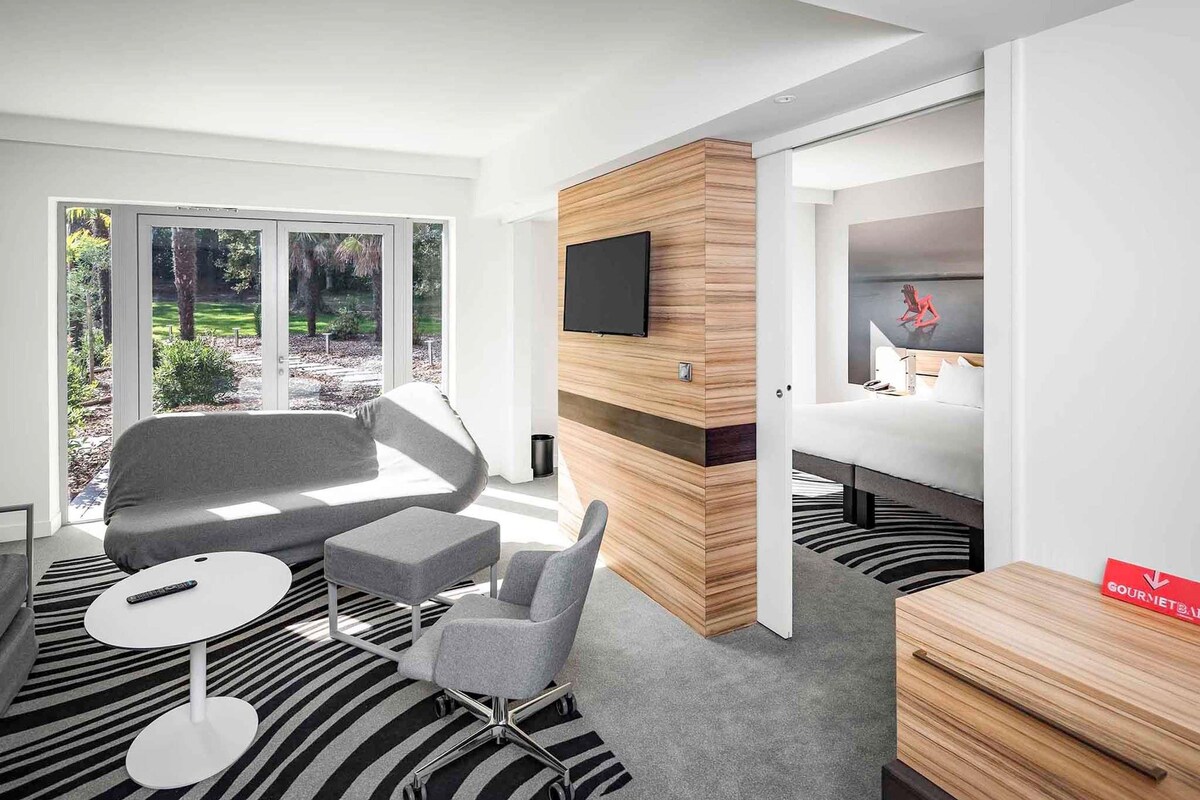 A modern hotel room with a large window, a flat-screen TV, a bed, a desk, and a chair.