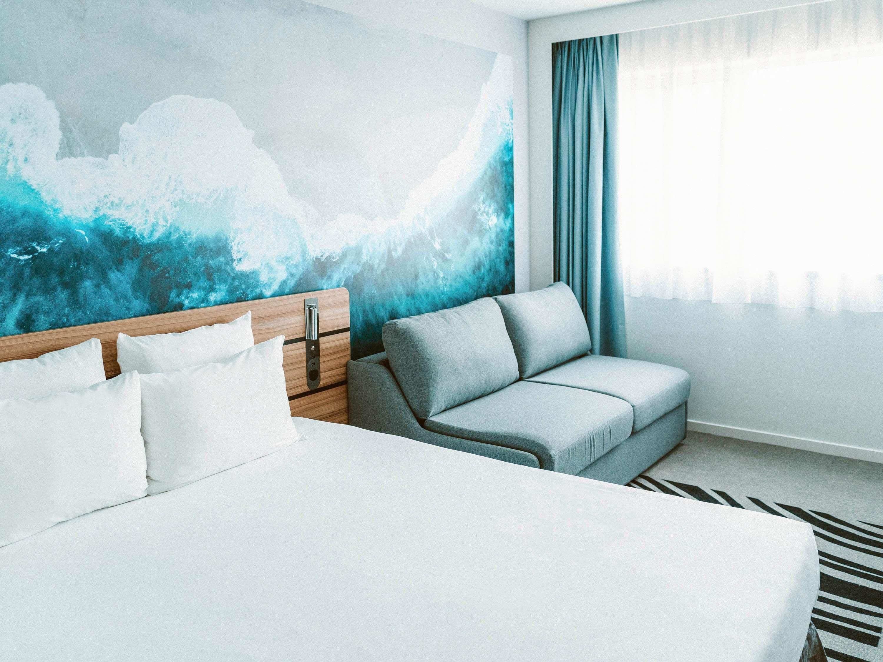 A hotel room with a bed, a grey sofa, a wooden headboard, and a blue abstract wall mural.