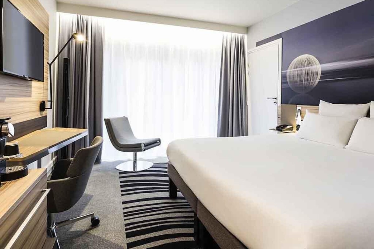A hotel room with a bed, a desk with a chair, a TV, and a large window with curtains.