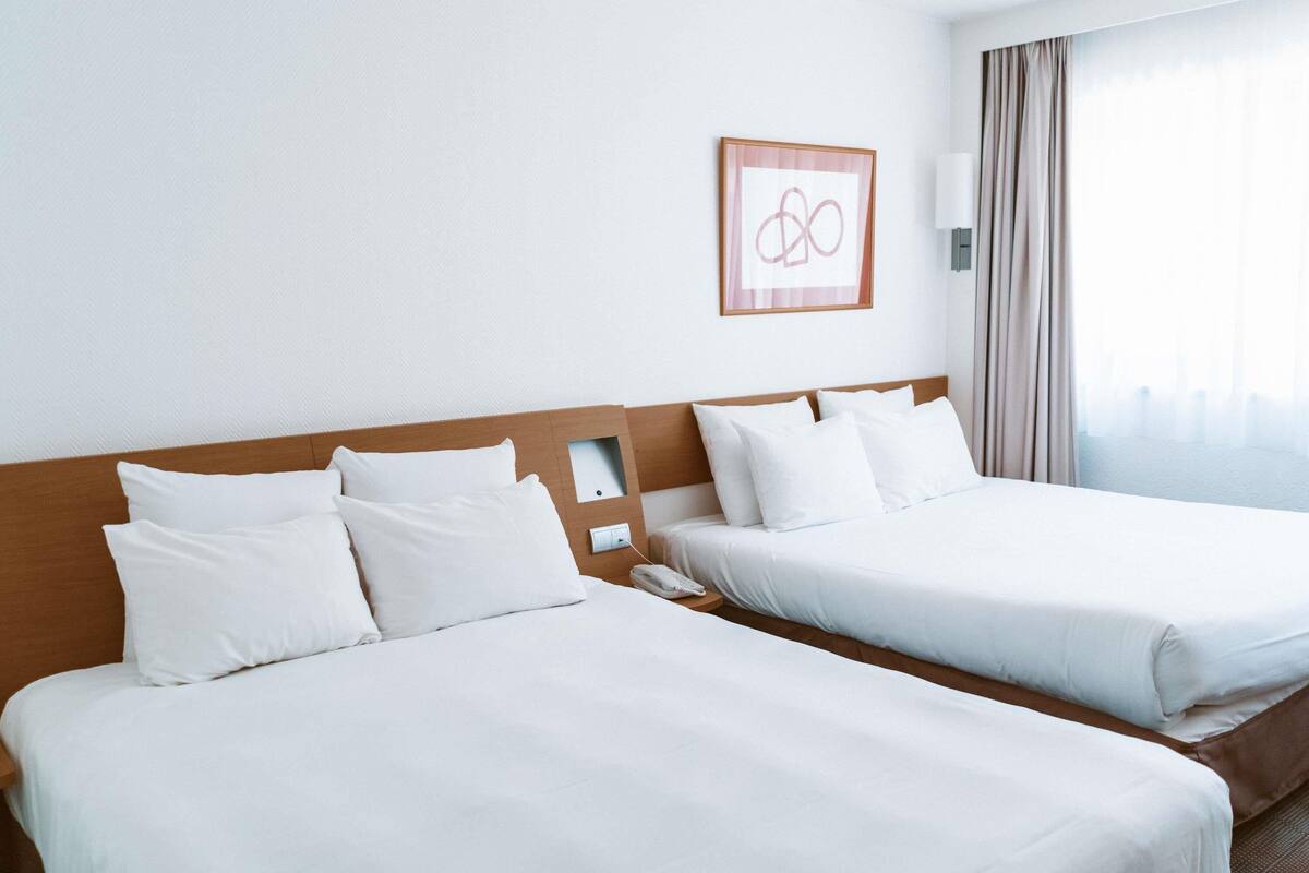 Dos camas individuales con cabeceros de madera, ropa de cama blanca, un cuadro enmarcado en la pared y una ventana con cortinas.