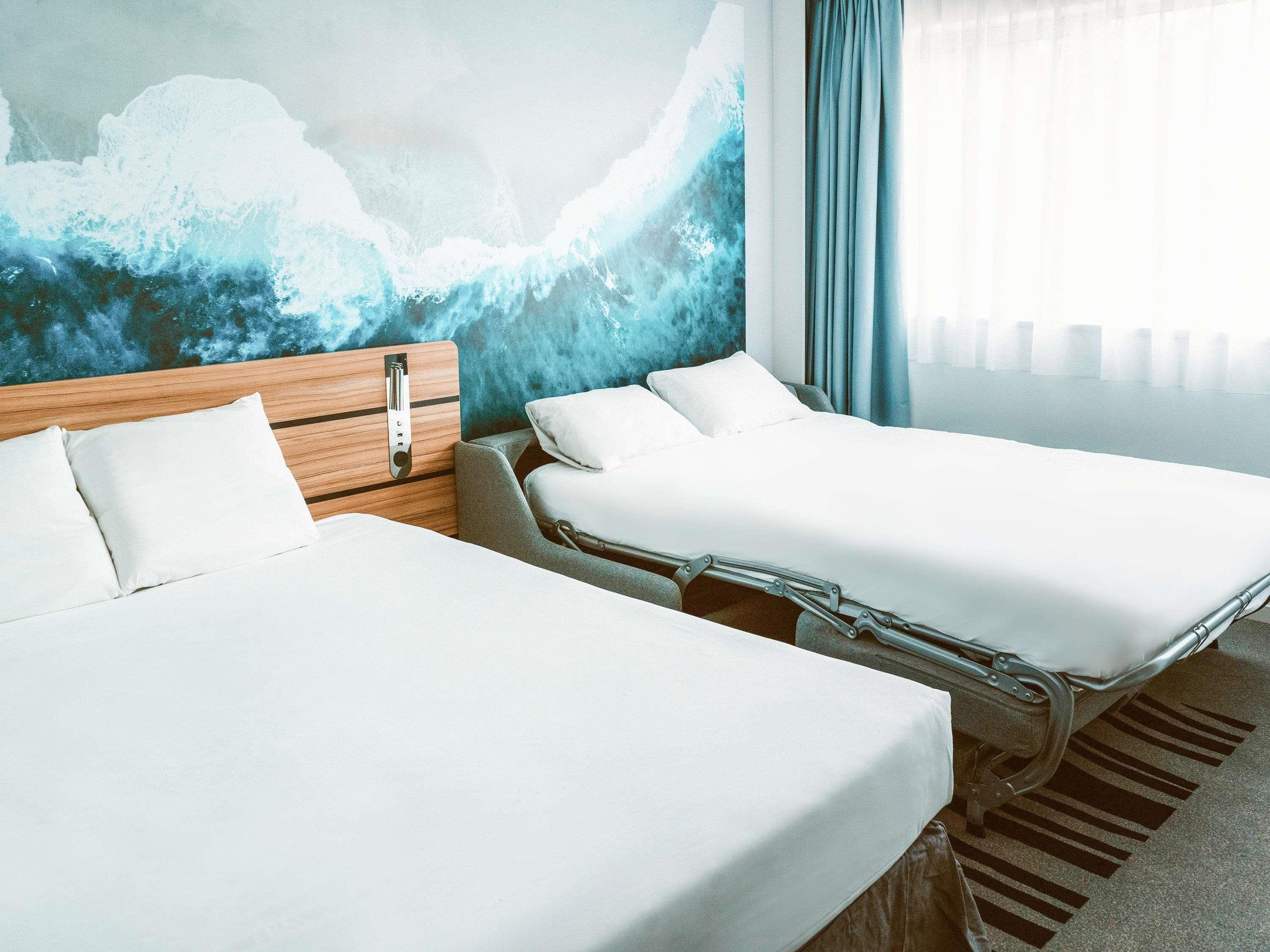 A hotel room with two beds, a wooden headboard, a large abstract mural, and a view of the sea.