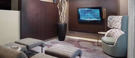 Una habitación de hotel moderna con un televisor de pantalla plana grande montado en la pared, un espacio de estar cómodo con sofá y otomanas, y una planta decorativa en una maceta negra.