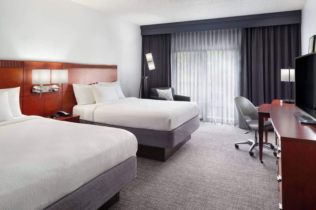 Een hotelkamer met twee bedden, een bureau, een stoel, een televisie en een groot raam met gordijnen.