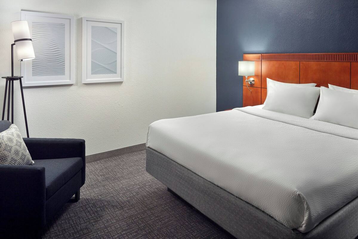 Een hotelkamer met een groot bed, een donkere fauteuil, een vloerlamp en twee kunstwerken in lijsten.