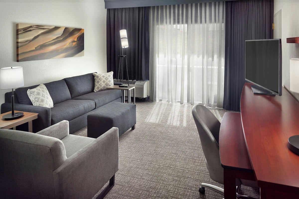 Een moderne hotelkamer met een grijze bank, een houten bureau, een flatscreen televisie en een groot raam met dunne gordijnen.