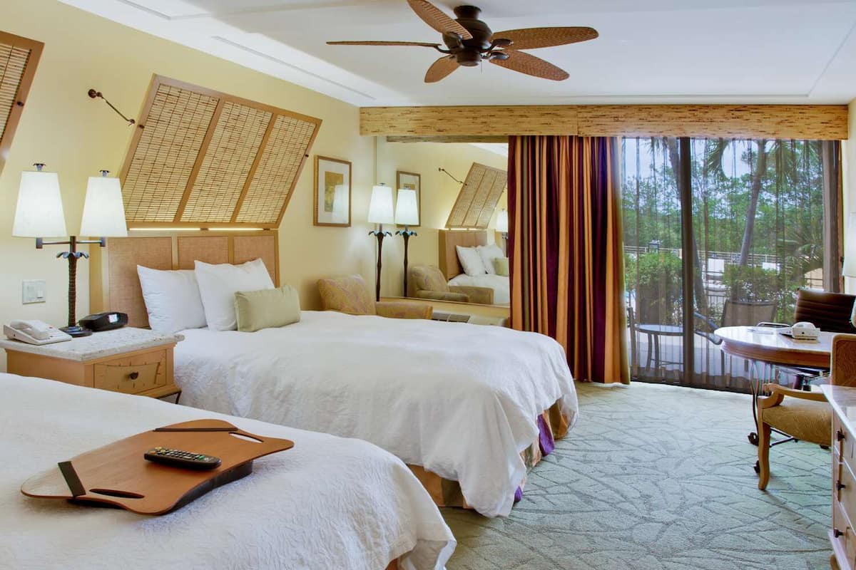A hotel room with two beds, a desk, a chair, a balcony, and a ceiling fan.