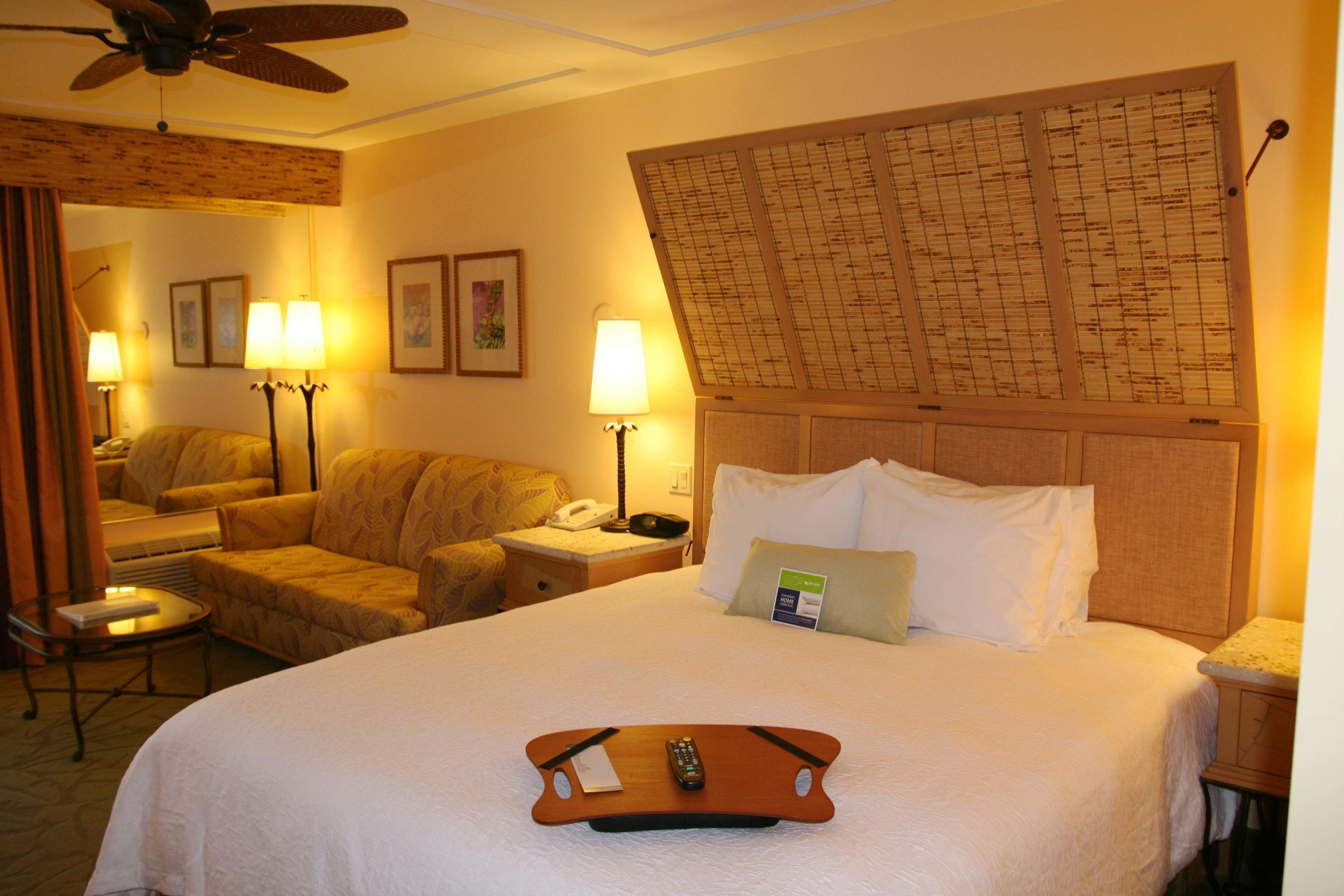 A hotel room with a large bed, a bedside table, a chair, a desk, a mirror, and a ceiling fan.