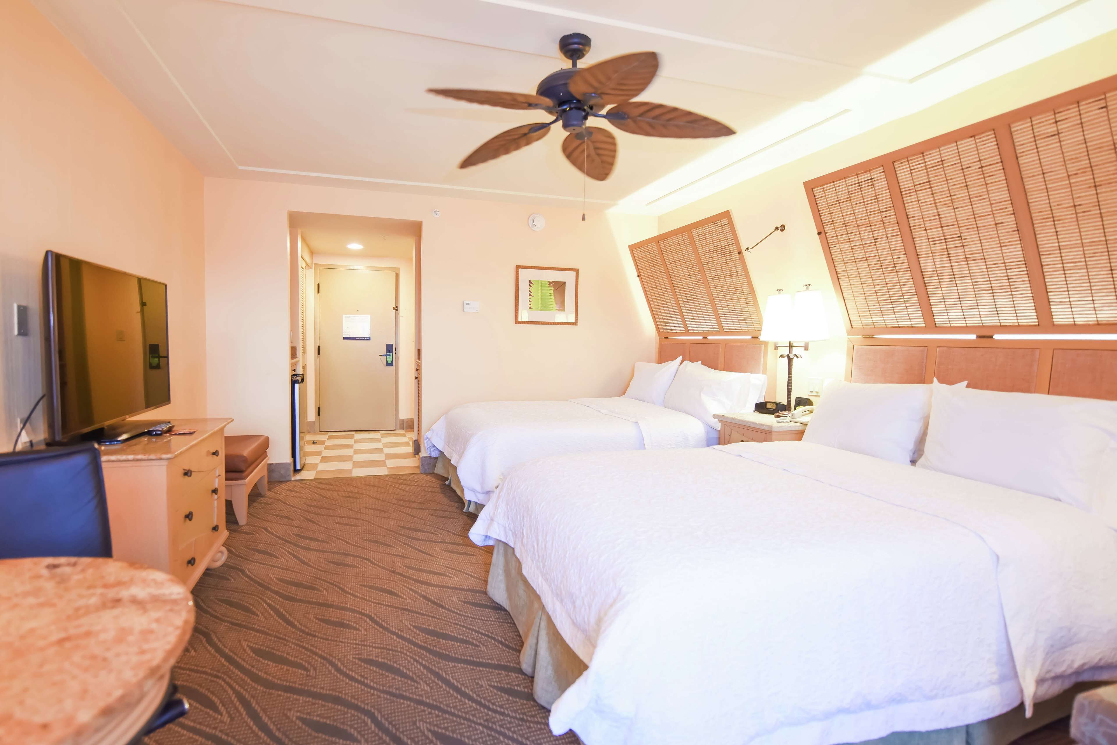 A hotel room with two beds, a ceiling fan, a TV, a desk, and a chair.