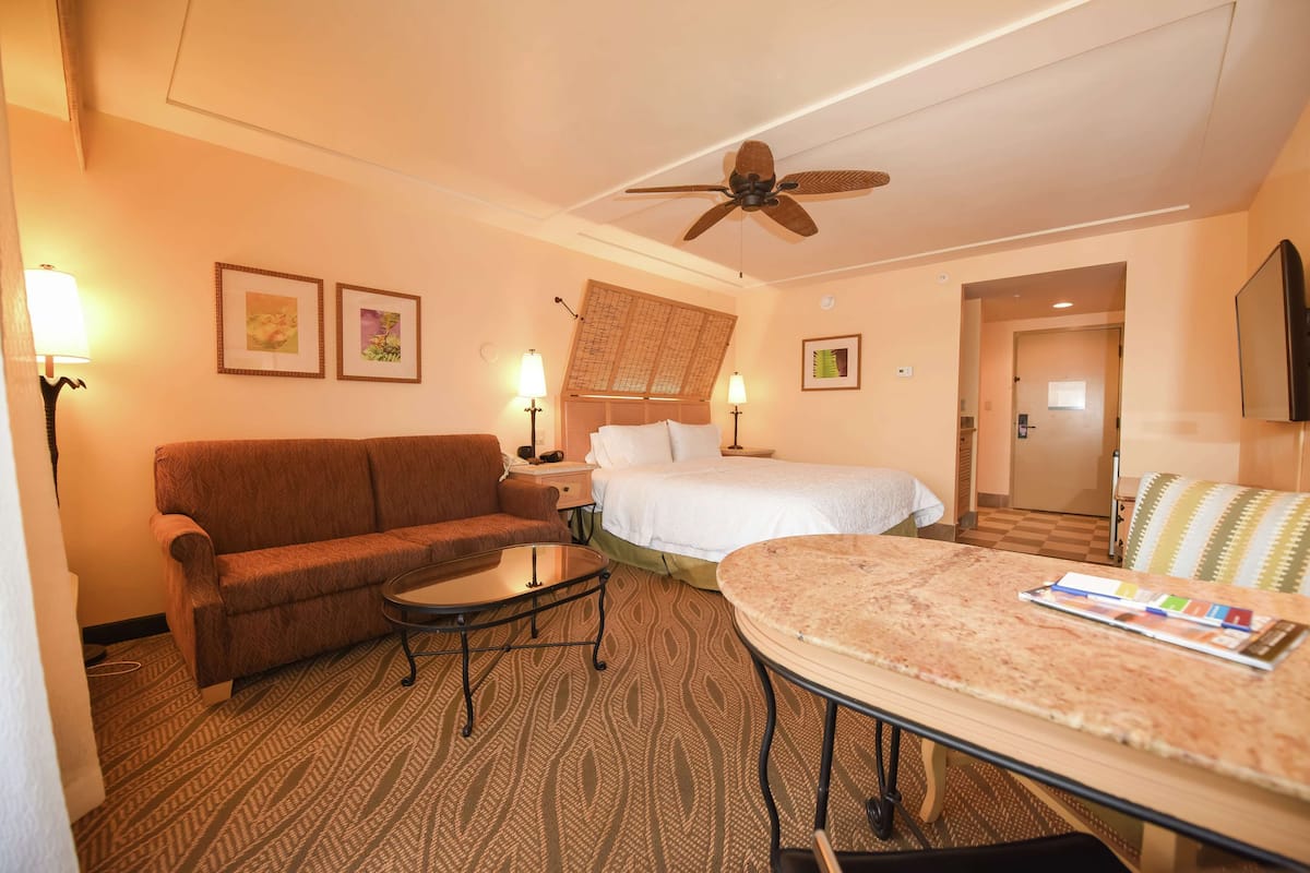 A hotel room with a bed, a sofa, a small table, a ceiling fan, and two wall-mounted lamps.