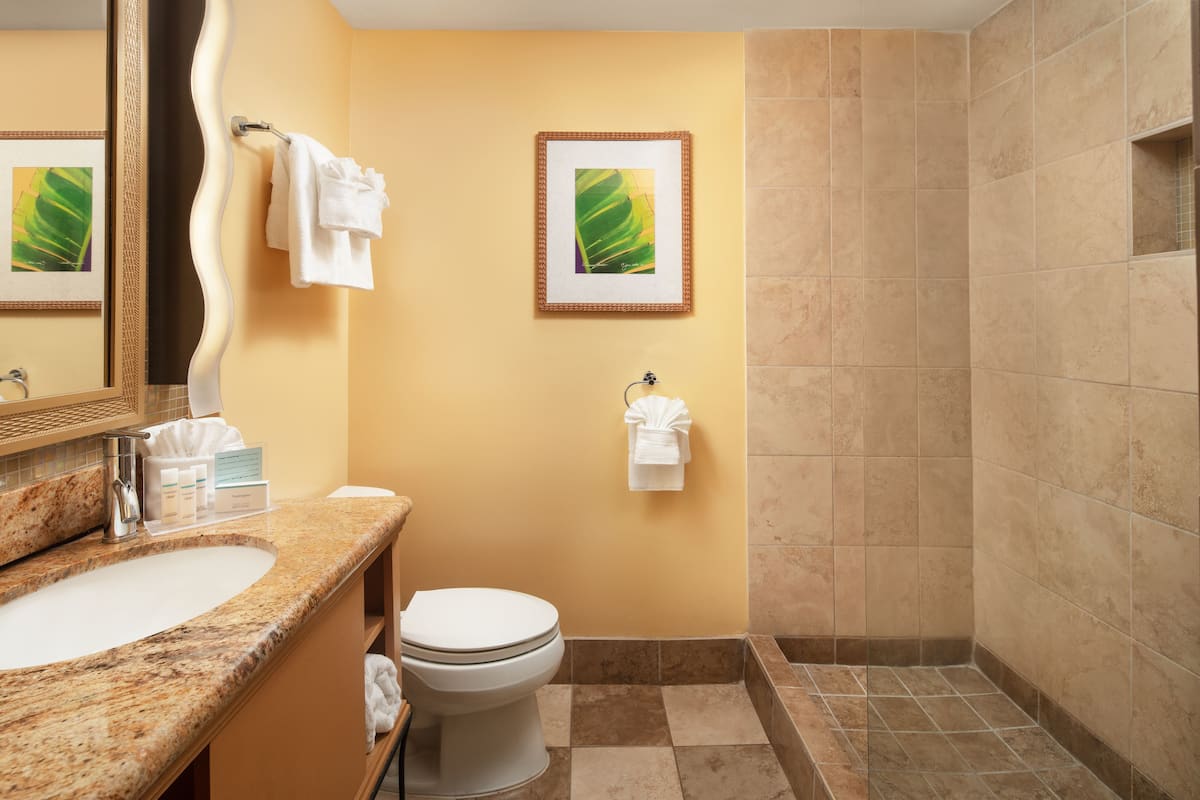 A bathroom with a sink, toilet, and shower. There are towels hanging on the wall and a framed picture above the toilet.