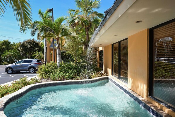 A swimming pool with a tiled edge, surrounded by lush greenery and palm trees, adjacent to a building with large windows and a parked car.