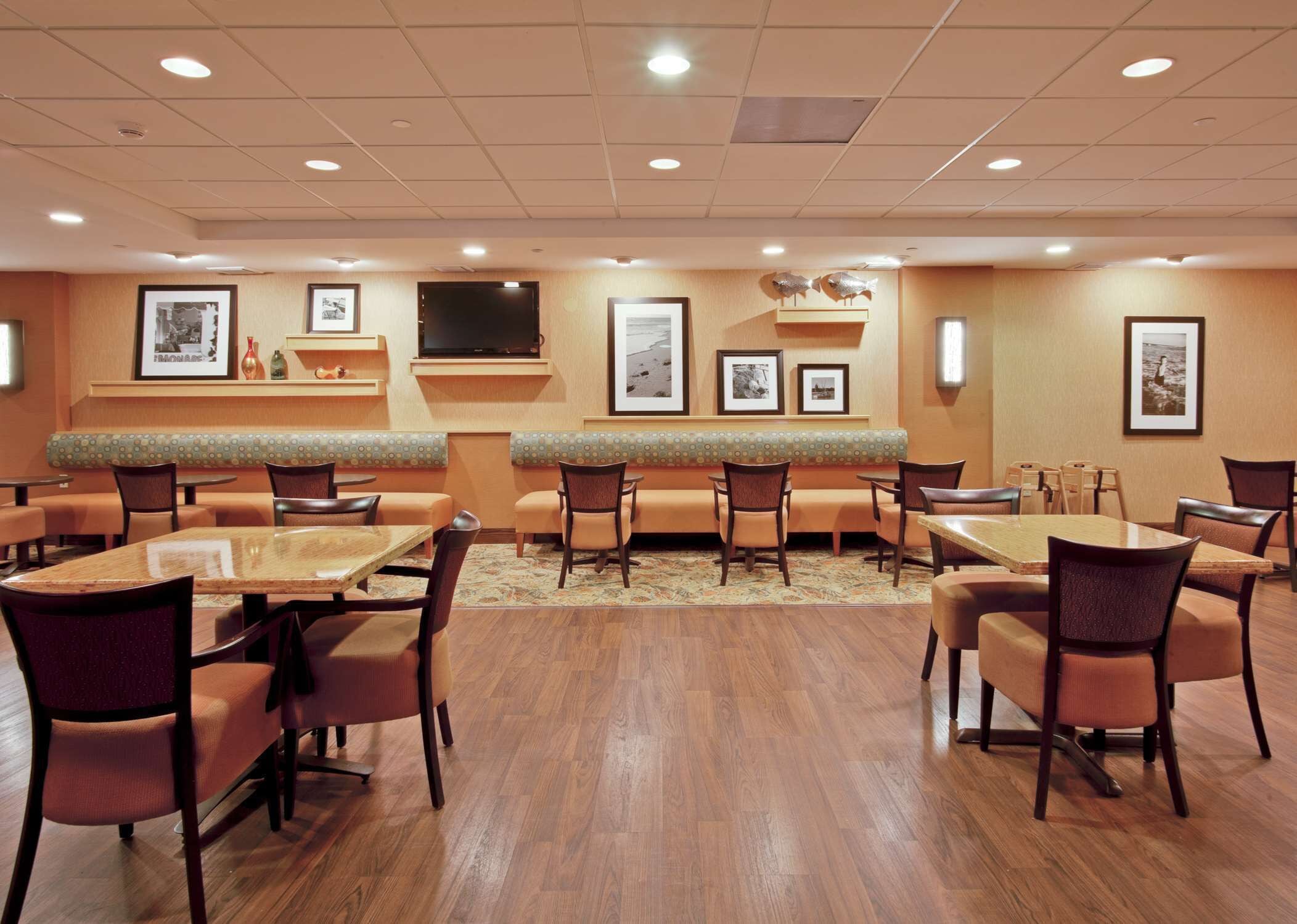 A hotel dining area with booths and tables, framed pictures on the walls, and a flat-screen TV above the booths.