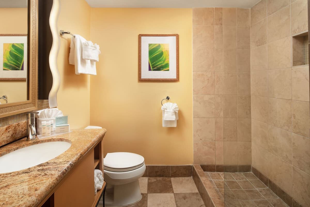 A bathroom with a sink, toilet, and shower. There are towels hanging on the wall and a framed picture above the toilet.